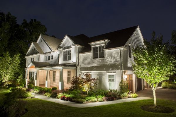 Front view of a large two-story house at night, illuminated by exterior lights. The yard is landscaped with plants and trees, and a concrete walkway leads to the entrance.